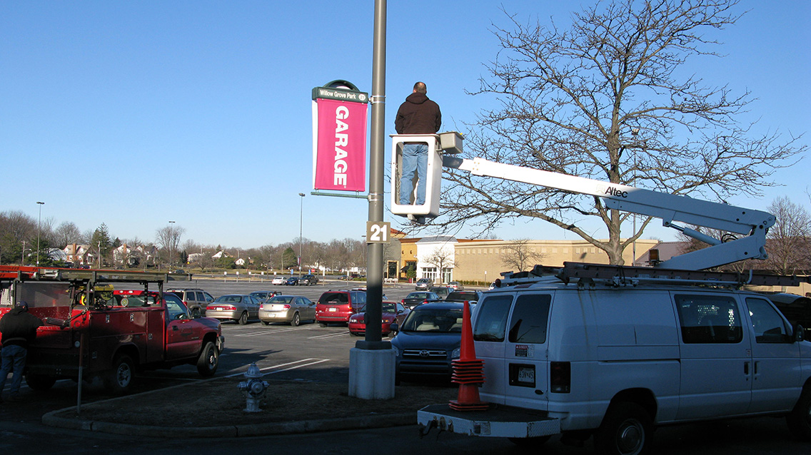 A Lumen Banner being installed on a light pole on in a parking lot by one man in a boom lift.